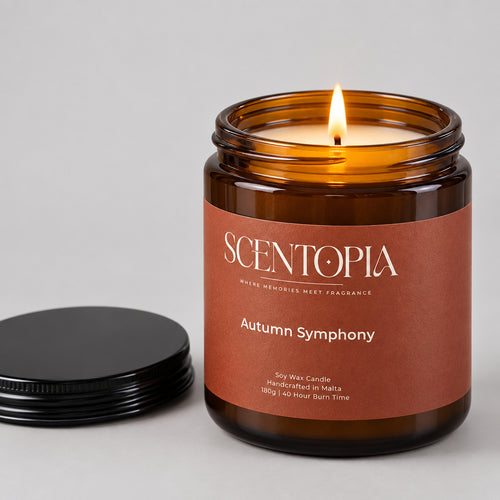 An amber glass jar candle labeled SCENTOPIA Autumn Symphony sits on a wooden surface with its black lid placed beside it against a plain beige background.