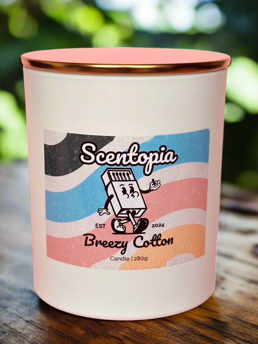 A white candle with a label that includes the brand 'Scentopia' and the scent 'Breezy Cotton'. The candle is on a pink background.
