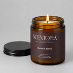 An amber glass jar candle with a dark brown label reading SCENTOPIA Burnout Blend, shown on a wooden surface with its black lid placed beside it against a plain beige wall.