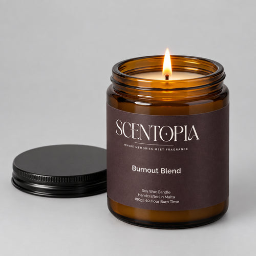 An amber glass jar candle with a dark brown label reading SCENTOPIA Burnout Blend, shown on a wooden surface with its black lid placed beside it against a plain beige wall.