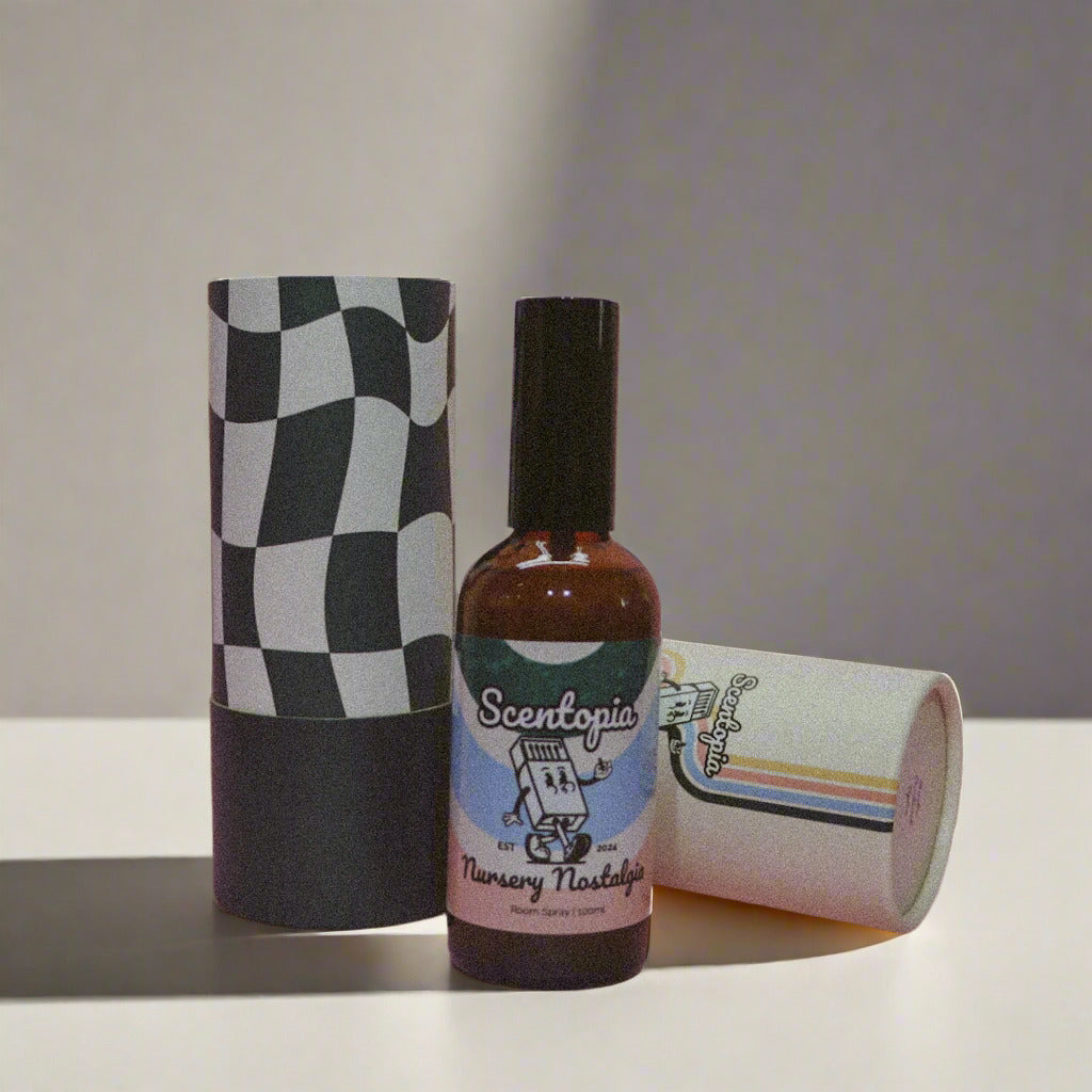 A bottle of 'Nursery Nostalgia' room spray by Scentopia, with a label featuring a vintage-style graphic. The bottle is placed in front of a checkered background, and there is a checkered box and a mug with a design in the background.