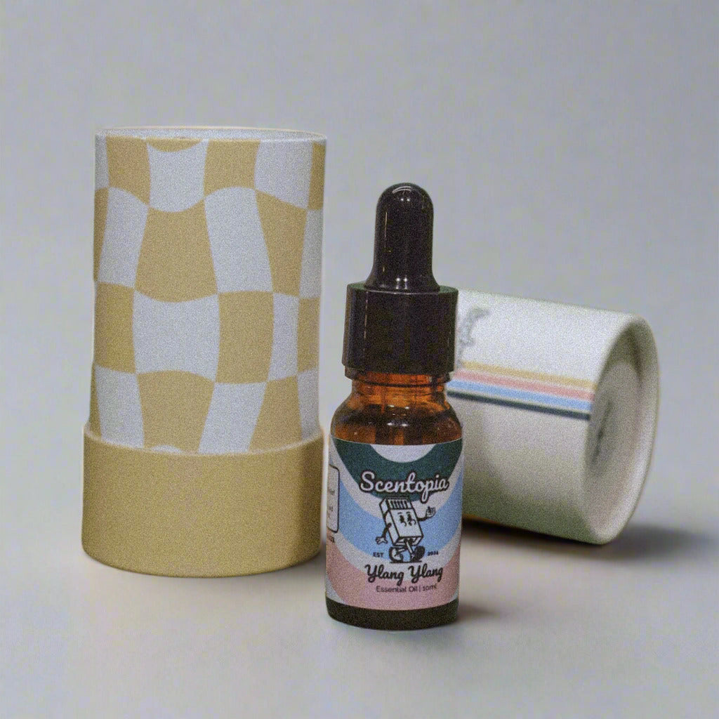 A 10ml amber dropper bottle of Scentopia Ylang Ylang essential oil stands in front of a tall cylindrical tube with a yellow and white retro checkerboard pattern, placed on a neutral surface.