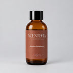 An amber glass bottle of Scentopia 'Autumn Symphony' room diffuser oil with a black cap, resting on a wooden surface against a neutral beige background.
