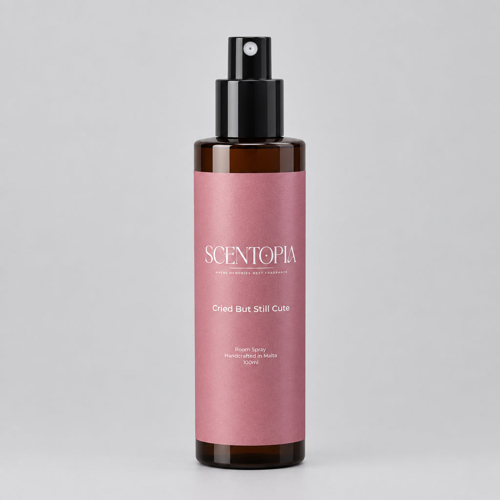 A bottle of Scentopia 'Cried But Still Cute' room spray, featuring a dusty pink label and a black spray nozzle, set on a wooden surface against a plain light background.