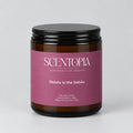 A Scentopia soy wax candle in a dark amber glass jar with a black lid and a dusty mauve-pink label, sitting on a wooden surface against a plain beige wall.