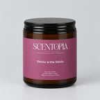 A Scentopia soy wax candle in a dark amber glass jar with a black lid and a dusty mauve-pink label, sitting on a wooden surface against a plain beige wall.