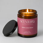 An amber glass jar candle with a pink label reading SCENTOPIA and 'Delulu is the Solulu,' shown on a wooden surface with its black lid placed beside it against a plain beige wall.