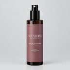 A bottle of SCENTOPLA Everyday Overthinker room spray, featuring a dusty rose label and black spray nozzle, standing on a wooden surface against a plain light gray wall.