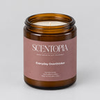An amber glass jar candle with a dusty rose label reading SCENTOPIA Everyday Overthinker, sitting on a wooden surface against a plain light background.