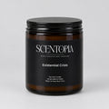 A SCENTOPIA soy wax candle in a dark amber glass jar with a black lid, labeled 'Existential Crisis', sitting on a wooden surface against a plain beige wall.