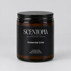 A SCENTOPIA soy wax candle in a dark amber glass jar with a black lid, labeled 'Existential Crisis', sitting on a wooden surface against a plain beige wall.