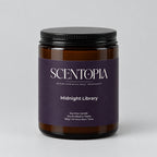 A Scentopia Midnight Library soy wax candle in a dark amber glass jar with a black lid and a dark purple label, sitting on a wooden surface against a plain beige wall.