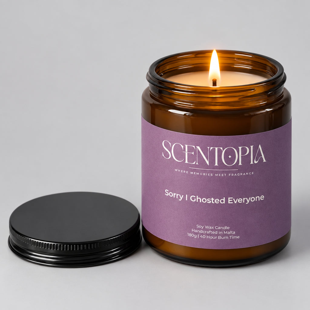 An amber glass jar candle with a lavender label reading SCENTOPIA Sorry I Ghosted Everyone, shown on a wooden surface with its black lid placed beside it against a plain beige wall.