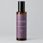A bottle of Scentopia 'Sorry I Ghosted Everyone' room spray, featuring a dark amber glass bottle, a black cap, and a lavender-purple label, set on a wooden surface against a plain light background.