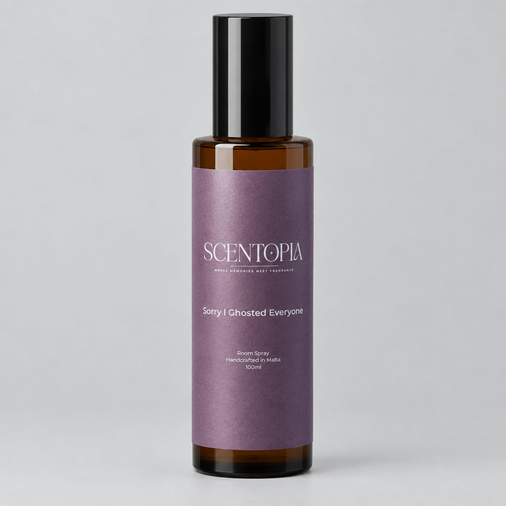 A bottle of Scentopia 'Sorry I Ghosted Everyone' room spray, featuring a dark amber glass bottle, a black cap, and a lavender-purple label, set on a wooden surface against a plain light background.