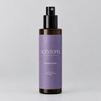 A bottle of Scentopia Wisteria Glow room spray with a lavender purple label and black spray nozzle, resting on a wooden surface against a plain light beige wall.